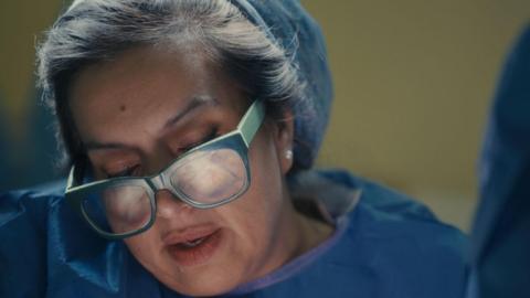 A close up of a woman with glasses and blue scrubs on, she is looking down and focusing on something