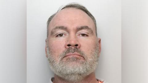 Wayne Stead with short dark hair and grey beard. He is looking at the camera in a police custody photo.