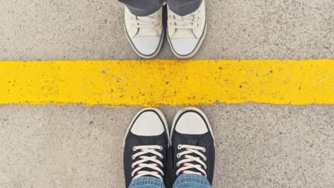 A yellow line on grey tarmac. Two pairs of shoes face each other on either side of the line. One pair is white and the other is black.