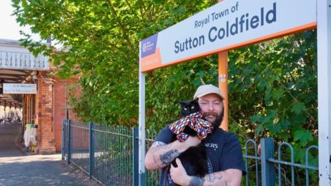 A man is standing on a train station platform holding a black cat. He has a cream cap on, black beard and tattoos. Behind him there is a sign saying "Royal Town of Sutton Coldfield", blue fence and platform. 