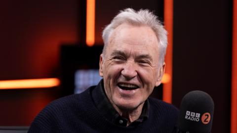 Larry Lamb pictures smiling at a BBC radio 2 microphone. He has spikey grey hair and is wearing a dark blue jumper with a dark green and blue checked shirt underneath.