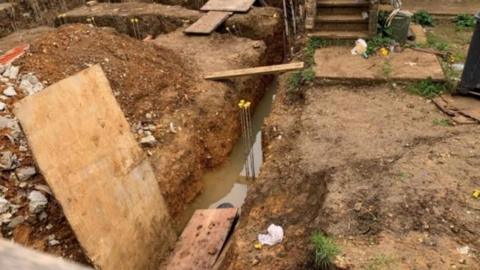 A long, narrow excavated trench running through the centre of a muddy brown construction site, with some muddy standing water visible at the bottom.