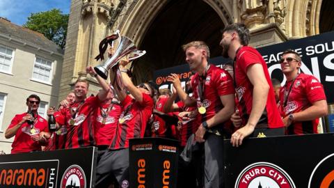 The team lift the trophy while standing on the steps of Truro Cathedral