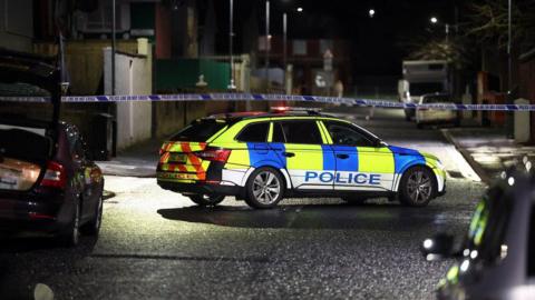 A blue and yellow police car is side on, on a street, facing the right side of the frame. There is blue and white police tape beyond the car, running parallel with the car. 
Two cars can be partially viewed in the foreground, on each side of the frame. 