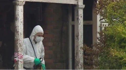 A man wearing white forensic overalls and green gloves emerges from a red brick building. 
