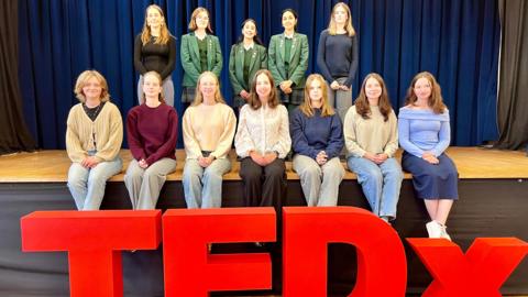 Twelve pupils from the Ladies' College in two rows on a stage - the front row of seven are sat and back row of five are standing - in front of a TEDx symbol.