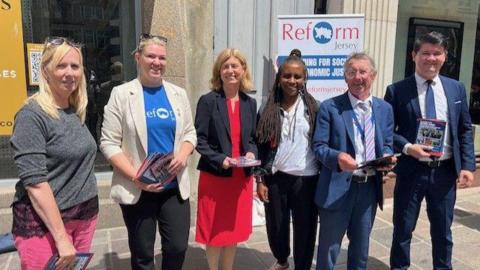 Six Reform Jersey members including Sam Mezec standing on a shopping street in a line holding leaflets and lookng at the camera 