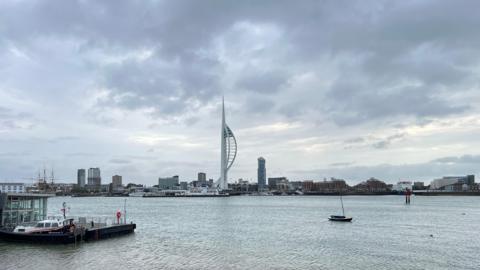 Portsmouth Harbour with grey skies overhead. The large whit Spinnaker tower is on the far side of the harbour where you can see the city skyline. On the left you can see the mast of HMS Warrior and there several smaller boats in the water. 