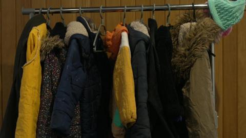 A clothes rack is full of coats with some yellow and blue ones standing out. There is a green hat hanging from the end of the rack and a brown coat with a furry hood next to it.