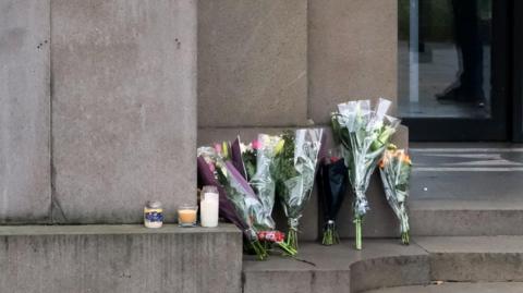 Flowers and candles at the Australian High Commission in London. There are several bunches of flowers and three candles at the top of stone steps.