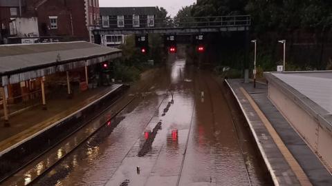 A flooded railway line, with brown flood water submerging train tracks next to two platforms.