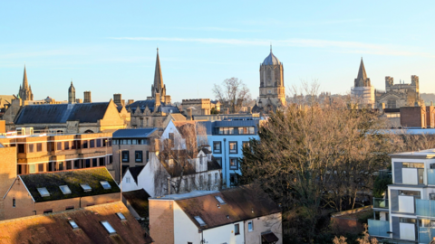 A city skyline under a clear blue sky on a winter's day. There are streets of homes and flats in the foreground with bare trees. In the distance you can see the spires of the Oxford colleges.