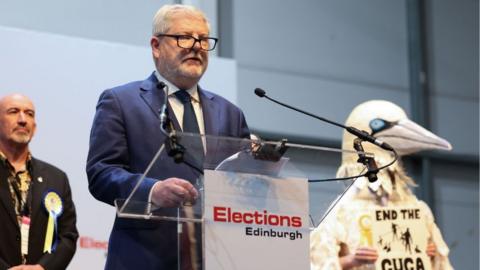 Angus Robertson, with grey hair and glasses, speaks at a podium, with a man dressed as a bird behind him 