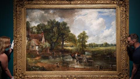 A large landscape oil painting in an ornate gilded frame is held in position on a wall by a woman to the left and a man to the right. The painting is The Hay Wain by John Constable, from 1821, and depicts a horse and cart in a shallow river, with a white house to the left. The scene is surrounded by trees and fields. 
