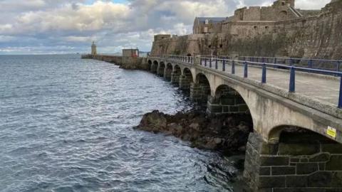 A long multi-arched bridge which leads to a lighthouse and a castle with the sea to its left.
