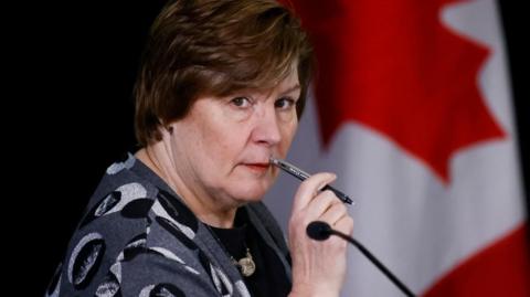 Commissioner Marie-Josée Hogue listens to hearings for independent commission probing alleged foreign interference in Canadian elections. She holds a pen up to her mouth. Behind her is a Canadian flag. Her expression is attentive. 