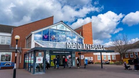 The entrance to a hospital. The sign above reads Main Entrance. People are pictured outside the entrance.
