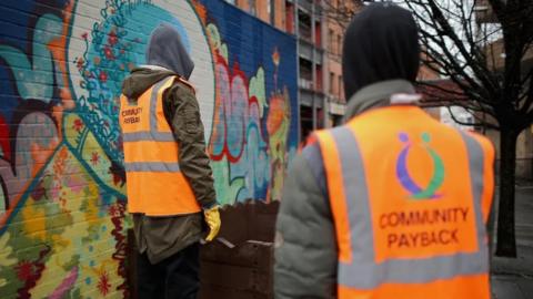 Two people wearing "community payback" orange hi-vis jackets looking at graffiti on a wall