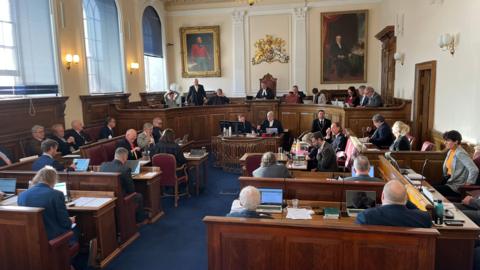 A government chamber with blue carpet and rows of wooden desks and chairs with various people in business attire sitting at them. A raised, curved court-style bench is at the back of the room with two oil painted portraits being and a royal crest.