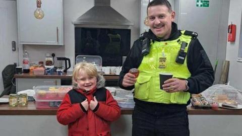 Alfie in a red coat smiling and standing next to a police officer who has bought a cake and coffee. Alfie is giving the thumbs and in the background of the kitchen there are iced buns in a tub. 