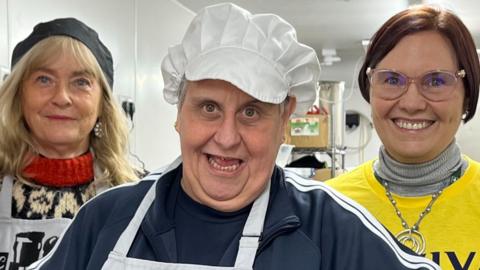 Three women stand in a kitchen. All are smiling. Two are wearing bakers' caps and aprons. 