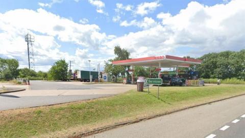 Google street view of Foston petrol station it has a red and white Esso canopy above the petrol pumps and shops and is surrounded by trees 