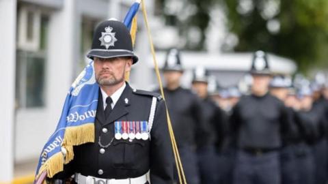 A police officer in black uniform with a row of medals and a tall helmet, carrying a blue flag with yellow tassels