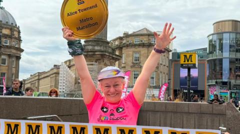 Alice Townend smiling into the camera with her arms up in the air. She is wearing a colourful cap with the brim standing up and has white hair peeking through. She is wearing a bright pink t-shirt and is holding a large cardboard medal in one of her hands which reads "Alice Townsend Metro Marathon". People are holding a white finishing line banner in front of her with the yellow Metro logo, which is a black capital M in a yellow box. The bottom of the stone Grey's Monument is behind her, as well as a large Metro sign. The stone buildings of Grainger Street are also behind her along with the glass-fronted Eldon Square.
