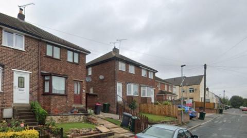 A street view of the row of houses in Clement Street. The houses are made from red brick, there are flowers and hedges in front lawns as well as a number of wheelie bins.