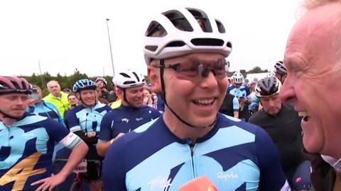 Sir Chris Hoy dressed in cycling gear, smiling and surrounded by other cyclists