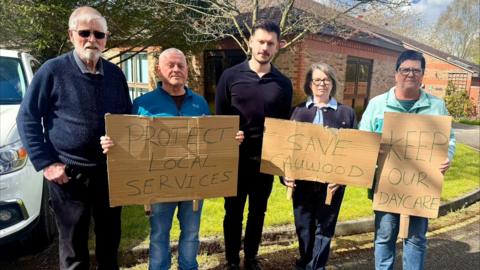 Cllr Keane Duncan is pictured standing in the centre of four other people. Three are holding signs saying 'Protect local services', 'Save Cauwood', and 'Keep our daycare'. Keane has brown hair and is wearing a navy blue shirt and dark trousers. Parent Caroline Garrod is to his right, with dark hair and wearing a light blue shirt with a navy jacket and dark trousers. Three other parents are in the photo, one a woman with short dark hair and glasses wearing a turquoise jacket and jeans, one a grey haired man wearing a bright blue jumper and jeans, and the final one a grey haired man wearing sunglasses, a navy blue jumper and dark trousers. They're standing in front of a brick building, with grass and a couple of trees behind them. There is also a white car to the left of the image.
