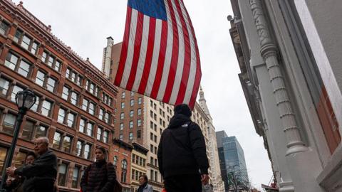 A street in the US with pedestrians walking around and a US flag hanging from a building.