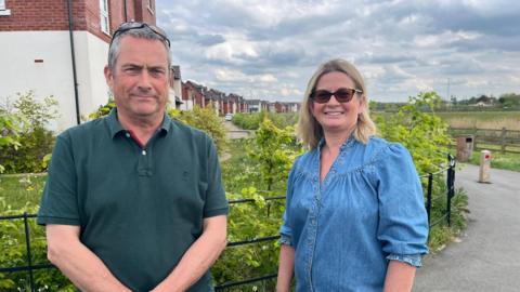 A man on the left wears a green polo neck, he has grey hair and sunglasses on his head. The woman wears a blue denim top and sunglasses, they are stood in front of a new build housing estate.