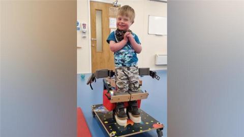 A young boy with dark blonde hair is wearing a blue camo print t-shirt and grey camo-print trousers. He is standing in a specialised standing frame which is on wheels, with his feet on blocks. He has wooden blocks in front of his knees and there is a waist strap which is undone behind him.