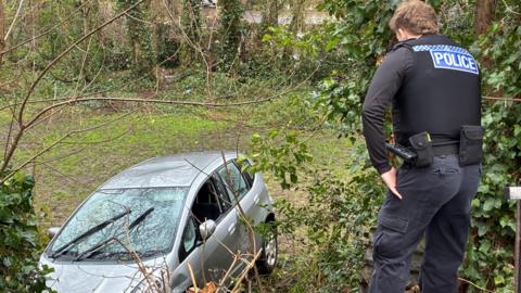 Silver car down an embankment with police officer standing on the bank with their back to the camera.