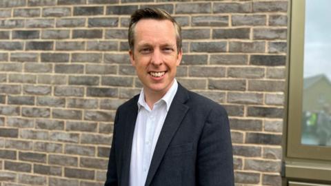 Tom Pursglove with short fair hair wearing a dark jacket and  white shirt in front of a brick wall.