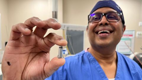 A doctor wearing blue surgical clothing and hat smiles as he hold a tiny pacemaker up to the camera.