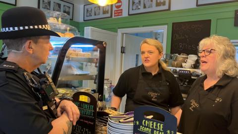PC Pip Davey is dressed in full uniform standing in front of a shop counter talking with Olivia and Jo Richards who are dressed in a shop uniform. There are coffee cups stacked behind them and a counter display with sandwiches and cakes and small blackboards detailing special offers including a cheese scone deal.