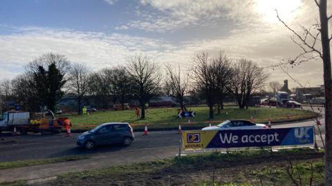 Roundabout with green grass in the middle with cars going around it. There is cones and a orange tape with roadworks ongoing.