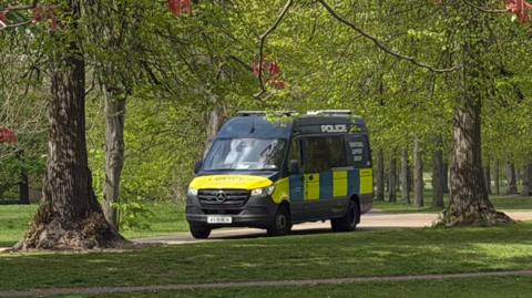 A yellow and blue police van is parked on a path among trees in Kensington Gardens