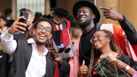 Stormzy is stood in the middle of two students. The man on the left is holding a phone taking a selfie. They are all looking towards the camera smiling. 