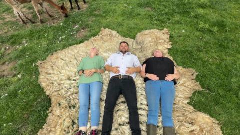 Sheep shearer Carl Sutton lies on the wool, alongside Lynn Kirk (left) and Angie Beale from the Black Barn Therapy Farm. The three of them are laying down on the fleece. It is easily taller than all three and much wider.