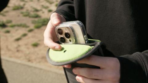 A person's hands placing a smartphone with a clear case into a small green fabric pouch. The scene is outdoors, with a blurred sandy and grassy background visible behind the hands.