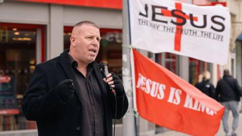 Preacher Stephen Clayden preaching on the street. There are flags reading 'Jesus, the way the truth the life' and 'Jesus is King'. 