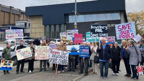 A group of men and women, outside a large building, holding placards with messages campaigning for the reopening of the leisure centre. The closed building can be seen in the background.  