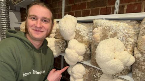 Kieran Sockett, a man in his early 30s with long hair and sideburns, knelt next to and pointing towards mushrooms blooming from bags of feed.
