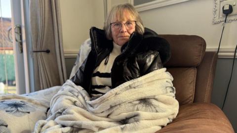 Jean Hull in a brown arm chair with a white long blanket covering her legs and arms.