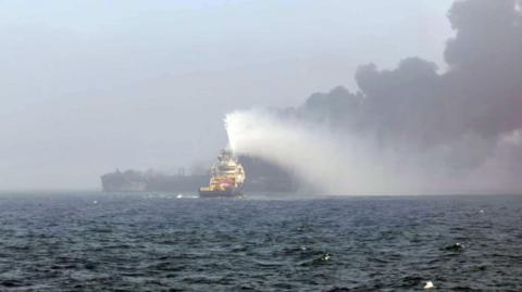 The image shows a boat on the sea with black smoke coming off it. There is a rescue boat in the foreground spraying water onto it