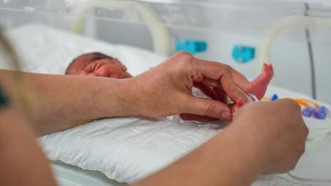 A tiny baby is seen laying in a neonatal intensive care bed. A pair of hands tends to the baby's foot. The infant lies on a white pad mattress.