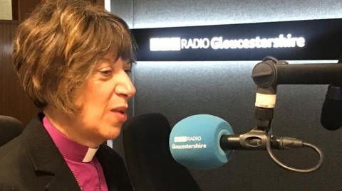 Right Reverend Rachel Treweek is sat in front of a BBC Radio Gloucestershire microphone in a BBC studio. A middle-aged woman with cropped brown hair.